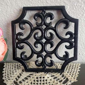May include: A black, square, cast iron trivet with an ornate, scrolled design. The trivet sits on a cream-colored, crocheted doily. The trivet's design is symmetrical and features swirling patterns.