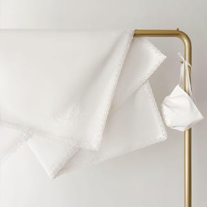 Op de afbeelding: Witte beddengoedset met geborduurde details en kanten rand, gepresenteerd op een goudkleurig rek. Een bijpassende witte tas hangt aan het rek. Het beddengoed lijkt gemaakt van een zachte, hoogwaardige stof.