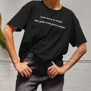 May include: Black t-shirt featuring the white text: "i just want to drink don julio with john summit". The top is a crew neck design and appears to be made from a soft fabric.