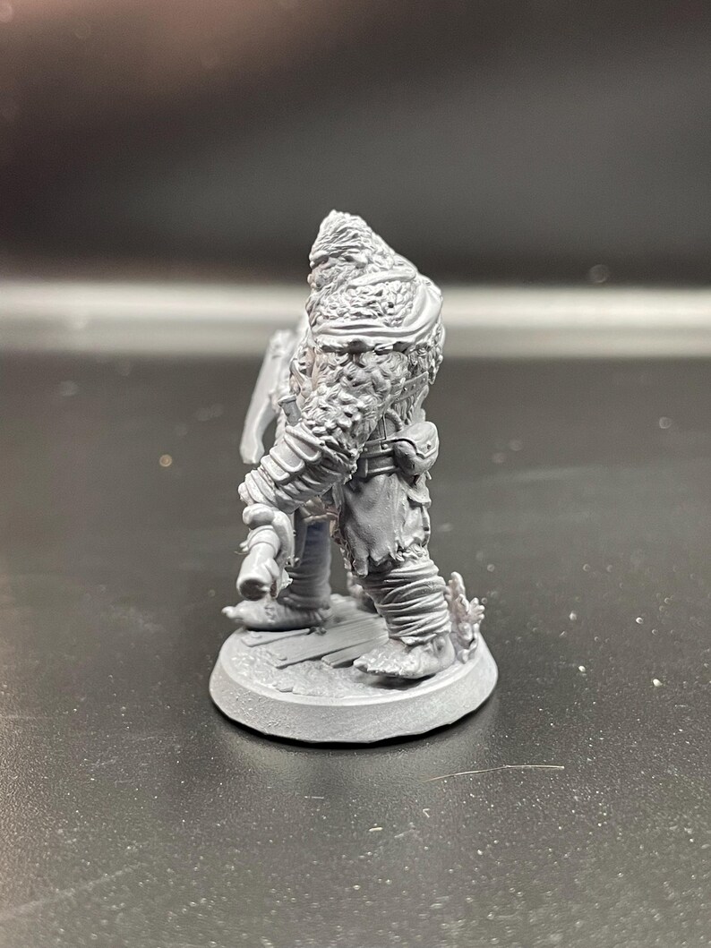 May include: A gray, unpainted miniature figurine of a furry, humanoid creature holding an axe. The figure is standing on a round base.