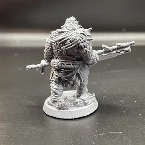 May include: A gray plastic miniature figurine of a fantasy creature with a large weapon. The creature is wearing a fur cloak and has a belt around its waist. The figurine is standing on a round base.