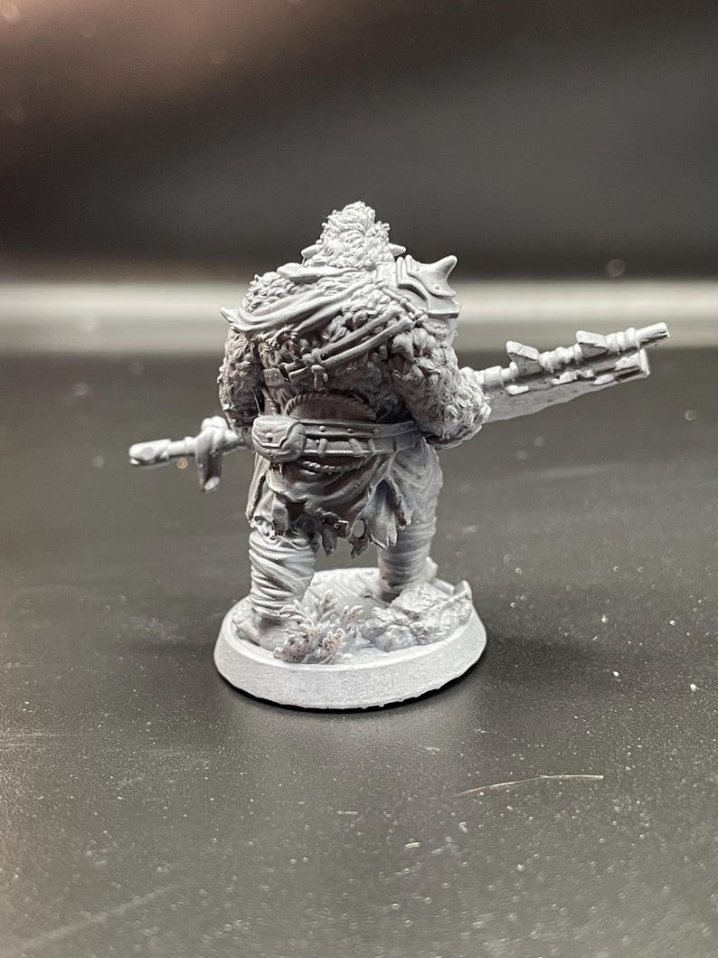 May include: A gray plastic miniature figurine of a fantasy creature with a large weapon. The creature is wearing a fur cloak and has a belt around its waist. The figurine is standing on a round base.