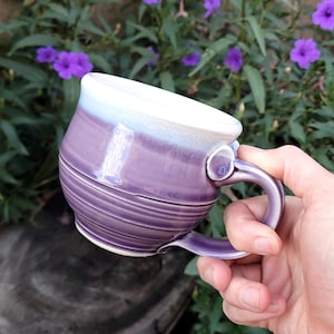 May include: A handcrafted ceramic mug with a purple and white glaze. The mug has a rounded shape with a handle and a textured, ridged exterior. The mug is held in a person's hand, with purple flowers in the background.