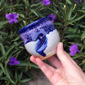 May include: A small ceramic cup with a rounded shape, featuring a speckled deep blue and purple glaze on the upper portion that transitions into a textured, off-white speckled base with a visible drip of blue glaze running down the side.