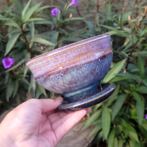 May include: A handmade ceramic bowl with a pedestal base. The bowl features a gradient of colors, including shades of blue, purple, and brown. The bowl is held in front of a background of green foliage and purple flowers.
