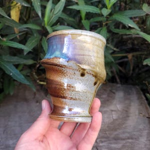 May include: Handmade ceramic cup with a unique, textured design. The cup features a blend of earthy tones, including brown, tan, and blue hues. The cup has a flared top and a slightly curved body, giving it a rustic and artistic appeal. The cup is approximately 4 inches tall.