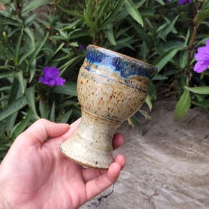 May include: A ceramic goblet featuring a speckled beige and tan body with a smooth, glossy deep blue glaze around the rim. The vessel has a wide, flared bowl that tapers down to a narrow stem and a circular base, showcasing a handcrafted texture.
