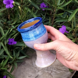 May include: A ceramic goblet featuring a flared rim and a stem base. The upper portion of the vessel is finished in a deep blue glaze, while the lower section and stem display a speckled light lavender and cream finish with a brown interior.