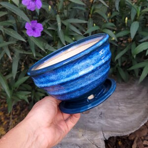 May include: A blue ceramic planter with a matching saucer. The planter has a glossy finish with horizontal ridges and a light interior. The saucer is attached to the bottom. The planter is held in a hand.