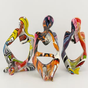 May include: Three colourful abstract sculptures of people sitting with their legs crossed. The sculptures are made of wood and painted in a variety of bright colours, including red, yellow, blue, green, and orange.