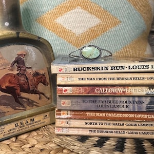 May include: A stack of paperback books with titles by Louis L'Amour. The books are all in a similar style with a brown spine and white text. The top book is titled "Buckskin Run". A turquoise and silver bracelet with a turquoise stone is on top of the books.