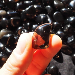 Peut inclure: Un ensemble de pierres d'obsidienne noires polies. Une pierre est tenue, mettant en valeur sa surface brillante et réfléchissante. Les pierres varient en taille et en forme, avec un aspect lisse et arrondi. Le fond est un tissu sombre et texturé.