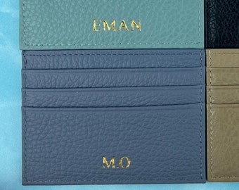 May include: Four leather cardholders in shades of blue, black, and tan, each with a different set of gold initials. The cardholders are arranged on a blue surface with floral accents. The cardholders have multiple card slots.