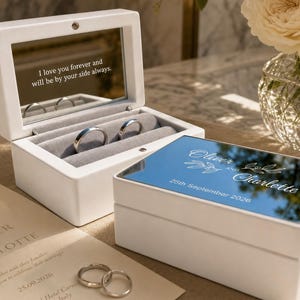 May include: A white wooden ring box with a mirrored lid featuring the engraved names Oliver and Charlette and the date 25th September 2026. The open box reveals a grey velvet interior with two silver rings and a mirror inscribed with I love you forever.