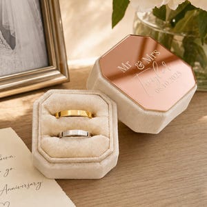 May include: A beige velvet ring box with an octagonal shape and a rose gold mirrored lid engraved with "Mr & Mrs Taylor 08.10.2025". The box contains two plain metal bands, one gold and one silver, resting in the velvet slots of the open base.