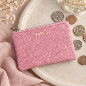 Può includere: Un portamonete rettangolare rosa con una texture granulata e una chiusura a zip color argento. Il nome JANET è impresso in lettere dorate al centro della parte anteriore. L'oggetto è posizionato su una superficie chiara con monete sparse.