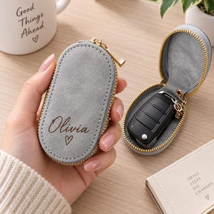 Könnte beinhalten: Ein ovales Schlüsseletui aus grauem Wildleder mit goldenem Reißverschluss. Die Vorderseite ist mit dem Namen Olivia und einem kleinen Herzsymbol personalisiert. Ein zweites passendes offenes Etui enthält einen schwarzen Autoschlüssel.