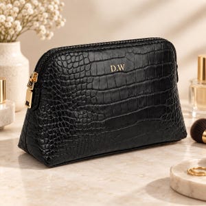 May include: A black cosmetic pouch with a textured crocodile skin pattern and a gold zip. The front of the pouch features the gold-coloured initials D.W. centred near the top. The bag has a structured, semi-circular shape with a flat base.