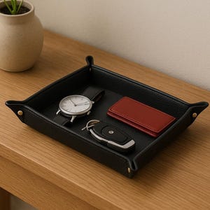Może przedstawiać: Czarna skórzana taca z zegarkiem, portfelem i kluczami na drewnianym stole. W pobliżu stoi mała roślina doniczkowa. Taca jest kwadratowa z zaokrąglonymi rogami i złotymi nitami.
