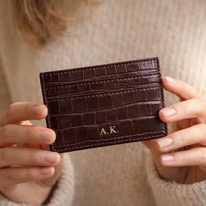 Op de afbeelding: Een bruinleren kaarthouder met krokodillenstructuur. De kaarthouder heeft meerdere sleuven en de gouden initialen "A.K.". De achtergrond is neutraal van kleur.