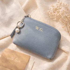 May include: A pale blue leather coin purse with a zip and gold initials "W.G.". It features a silver keyring with a pearl charm and a small leather tassel. A card reads "with love".