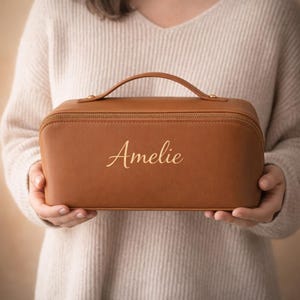 Puede incluir: Un neceser rectangular de color tostado con un asa superior y cierre de cremallera. La parte frontal presenta el nombre Amelie en una fuente cursiva. El neceser tiene un exterior liso de color sólido y es sostenido por dos manos.