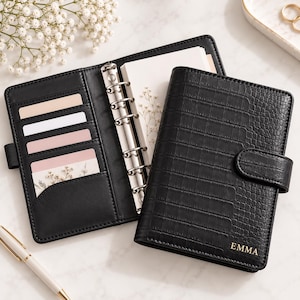 May include: A black faux leather personal planner with a crocodile texture and the name EMMA embossed in gold on the front cover. The open planner features a six-ring silver metal binder mechanism, multiple interior card slots, and white pages.
