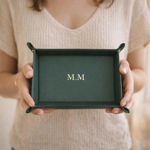 May include: A dark green leather tray with the initials "M.M" in gold lettering. The tray is rectangular with folded corners, designed for small items. The tray is held by a person.
