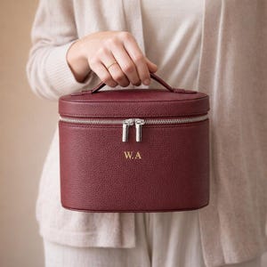 Puede incluir: Estuche de cosméticos burdeos con cremallera plateada y asa superior. La funda tiene las iniciales doradas "W.A". La funda cilíndrica parece ser de cuero.