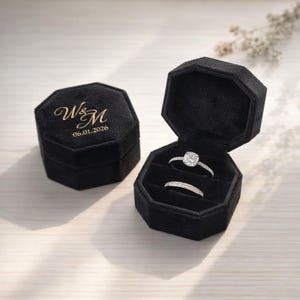 Puede incluir: Dos joyeros de terciopelo negro, uno abierto para revelar dos anillos. Los anillos son plateados con piedras transparentes. La caja cerrada tiene letras doradas y la fecha 06.01.2026. Los joyeros son octogonales.