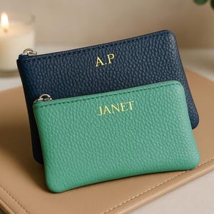 Puede incluir: Dos monederos de cuero personalizados, uno azul marino y otro verde azulado, con letras doradas. El monedero azul marino tiene "A.P" y el verde azulado tiene "JANET". Están sobre una superficie de cuero color beige, con una vela encendida y una planta en el fondo.