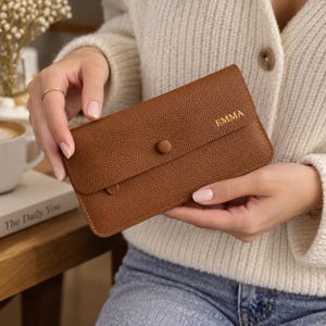 May include: A rectangular brown wallet with a textured surface and a single button closure. The name EMMA is embossed in gold lettering on the front flap. The wallet features a slim profile and a flap design that covers the main compartment.