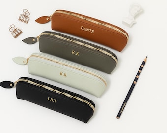 May include: Four personalized pencil cases in brown, olive green, pale green, and black, each with a gold zipper and a name or initials. A pencil, paper clips, and a small bust are also visible.