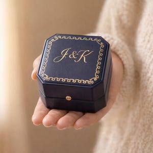 May include: A navy blue octagonal ring box featuring a gold-colored decorative border and the gold-colored script letters J&K printed on the lid. The box has a small gold-colored metal clasp on the front edge and a smooth, matte finish.