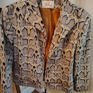 May include: A long-sleeve blazer featuring a tan, grey, and black snakeskin pattern. The jacket has a notched lapel, a single horizontal seam detail across the lower front, and a solid mustard-yellow lining. A label inside reads The Brittany.