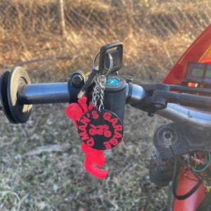 May include: A black and red keychain with the text "OLMAN'S GARAGE" and a motorcycle graphic. The keychain is attached to a set of keys and hangs from the handlebars of a motorcycle.