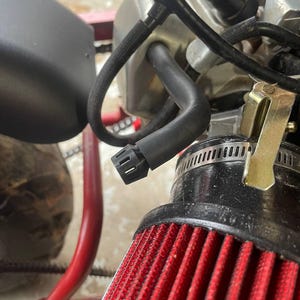 Op de afbeelding: Close-up van een rood luchtfilter met een zwarte behuizing en een zwarte slang eraan bevestigd. Het luchtfilter maakt deel uit van een kleine motor.