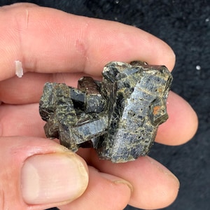 May include: A dark, rough-textured mineral specimen, primarily black with hints of green and brown. The crystalline structure is visible, with sharp edges and a slightly reflective surface. The mineral is held in a hand against a dark background.