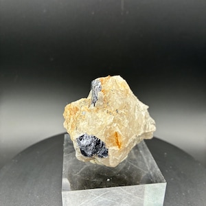 May include: A raw mineral specimen featuring a light tan and clear quartz matrix with embedded dark, metallic-looking crystals. The mineral sits on a clear, rectangular display stand against a dark background.