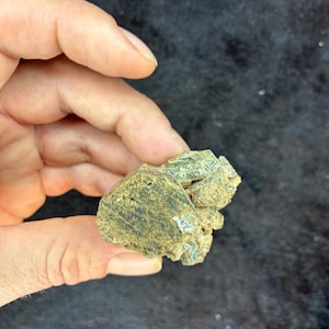 May include: A small, irregularly shaped mineral specimen with a mix of olive green and brown hues. The surface has a rough, textured appearance, with visible crystalline structures. The mineral is held in a hand against a dark background.