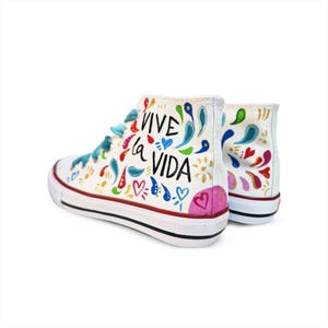 Op de afbeelding: Witte hoge sneakers met handgeschilderde ontwerpen. De sneakers hebben de tekst "Vive la Vida" in het zwart, met blauwe veters en een rode streep op de zool. Het ontwerp bevat harten en bloemmotieven.