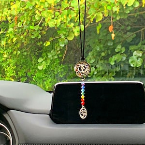 May include: A car charm featuring a silver-colored, filigree-style pendant with the Om symbol, hanging from a black cord. Below, a strand of colorful beads leads to a smaller Om symbol. The background shows a green, leafy environment.