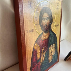 Może przedstawiać: Drewniana ikona z czerwoną ramą przedstawiająca Jezusa Chrystusa w złotej aureoli. Jest ubrany w czerwoną szatę na zielonej tunice i trzyma książkę. Tekst "IC XC NI KAITO&Rho;" jest napisany po grecku na ikonie.