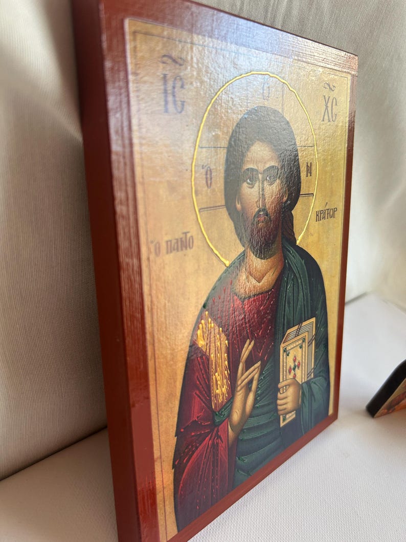 Może przedstawiać: Drewniana ikona z czerwoną ramą przedstawiająca Jezusa Chrystusa w złotej aureoli. Jest ubrany w czerwoną szatę na zielonej tunice i trzyma książkę. Tekst "IC XC NI KAITO&Rho;" jest napisany po grecku na ikonie.