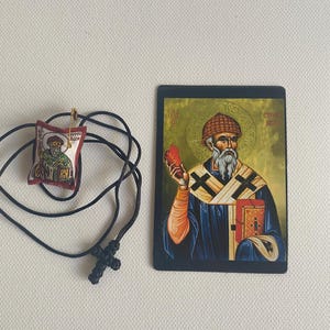May include: An Orthodox Christian icon and pendant necklace. The icon features a religious figure holding a book and a red object. The pendant is a small, embroidered image with a gold pin and a black cord necklace with a cross.