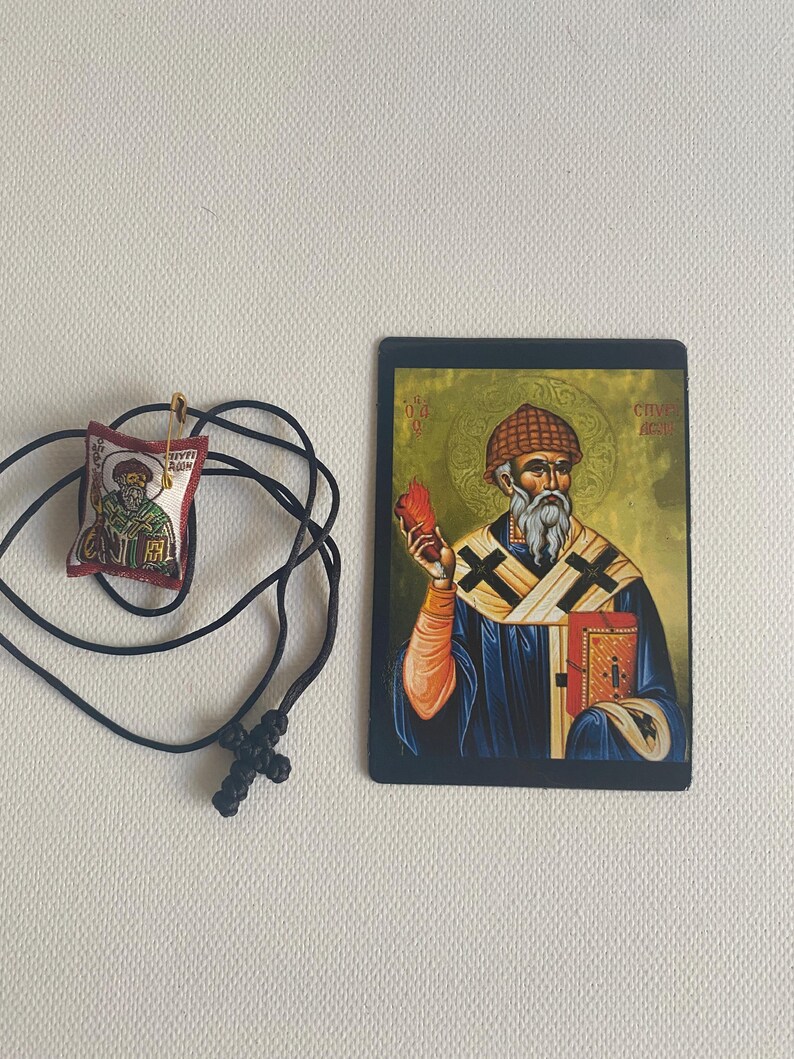 May include: An Orthodox Christian icon and pendant necklace. The icon features a religious figure holding a book and a red object. The pendant is a small, embroidered image with a gold pin and a black cord necklace with a cross.