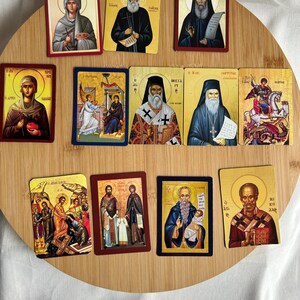 May include: A collection of rectangular religious icon cards featuring various saints and religious figures. The cards have gold, red, and blue borders, and the images depict figures in traditional religious attire. The cards are arranged on a round wooden surface.