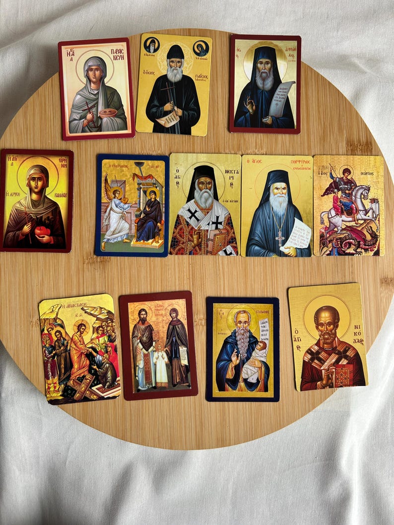 May include: A collection of rectangular religious icon cards featuring various saints and religious figures. The cards have gold, red, and blue borders, and the images depict figures in traditional religious attire. The cards are arranged on a round wooden surface.