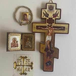 May include: A collection of religious items including a large wooden crucifix with a depiction of the crucifixion, a small folding wooden diptych icon, a circular beaded prayer rope with a small fabric icon, and a gold-coloured cross sticker with text.