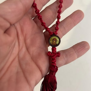 May include: A red knotted rosary with a tassel and a small circular pendant featuring an image. The rosary has a cross-shaped knot and is held in a hand.